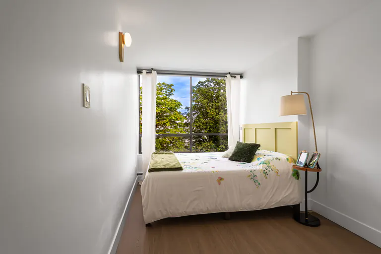 Modern narrow bedroom featuring a bed with green accents, a light green headboard, a large window with a tree view, and a brass floor lamp.