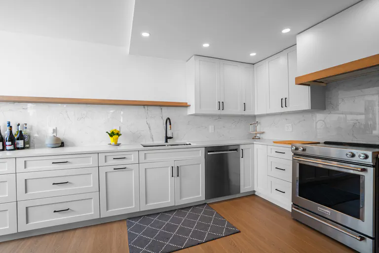 Modern white L-shaped kitchen with quartz countertops, stainless steel appliances, and wood floors.